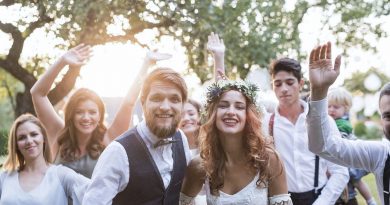 “Pareja vestida de boda sonriendo al frente mientras un grupo de invitados los rodea y celebra al aire libre, con árboles y luz de atardecer en el fondo.”