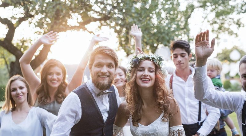 “Pareja vestida de boda sonriendo al frente mientras un grupo de invitados los rodea y celebra al aire libre, con árboles y luz de atardecer en el fondo.”
