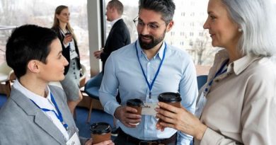 Tres personas con credenciales conversan mientras sostienen vasos de café en un espacio tipo oficina o sala de conferencias. Dos mujeres y un hombre están en primer plano, sonriendo y participando en una charla informal. Al fondo, otras dos personas también conversan. El ambiente parece ser un evento empresarial o de networking profesional.