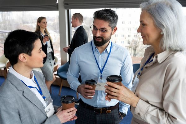 Tres personas con credenciales conversan mientras sostienen vasos de café en un espacio tipo oficina o sala de conferencias. Dos mujeres y un hombre están en primer plano, sonriendo y participando en una charla informal. Al fondo, otras dos personas también conversan. El ambiente parece ser un evento empresarial o de networking profesional.