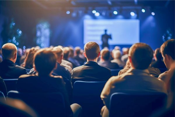 Un público numeroso sentado en un auditorio observa a un ponente que presenta frente a una pantalla iluminada. El ambiente es profesional, con iluminación tenue y enfoque en el escenario.