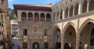 Fachada de un edificio histórico con arcos y balcones, situado en una plaza llena de gente, con banderas colgadas y ambiente festivo.
