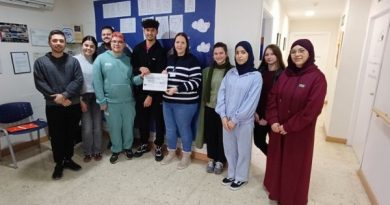Un grupo de jóvenes posa en un pasillo interior junto a una mujer adulta, sosteniendo un cheque. Están sonriendo frente a un mural azul con nubes y texto en la pared, vestidos con ropa informal y de pie en fila.