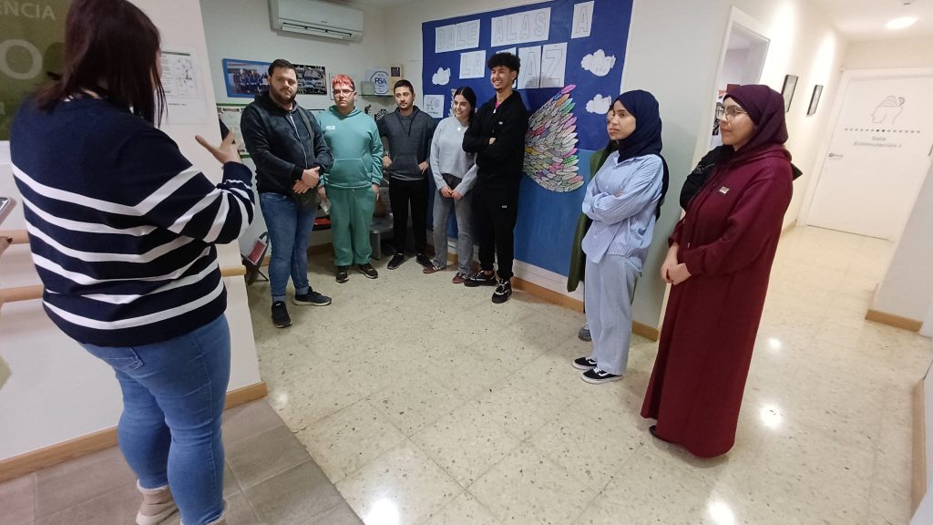 Un grupo de personas jóvenes y adultas escucha atentamente a una mujer que habla y gesticula en un pasillo interior. Están colocados en semicírculo frente a un mural azul con el mensaje “Dale alas a la paz” y unas alas de colores. El ambiente parece educativo o comunitario, con actitud de atención y diálogo.

