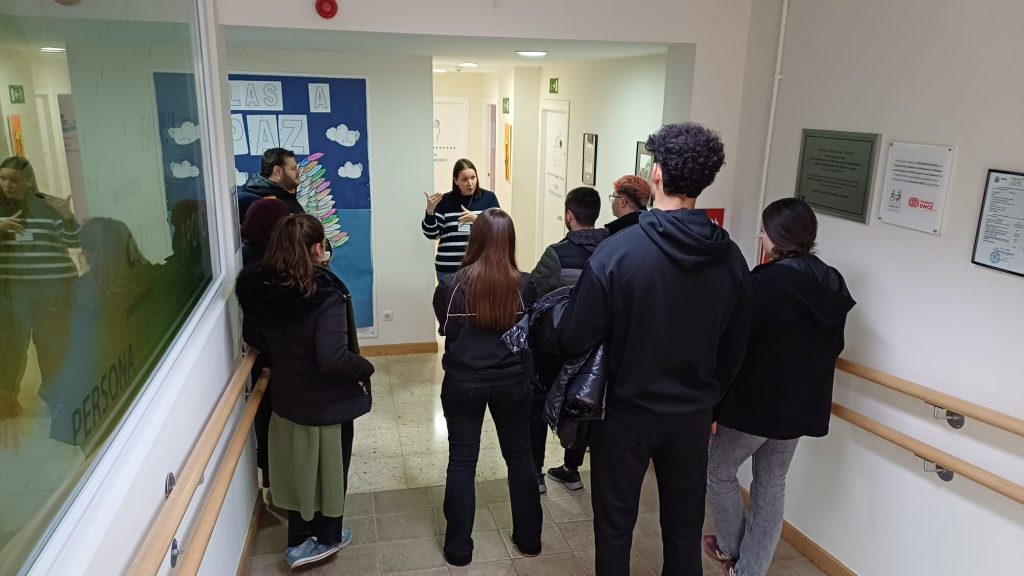 Grupo de personas adultas de pie en un pasillo interior de una asociación, atendiendo a una mujer que les habla y gesticula mientras explica algo. El pasillo es luminoso, con barandillas en las paredes y carteles informativos visibles. El grupo parece estar realizando una visita guiada o recibiendo indicaciones dentro del centro.