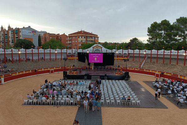 Vista general de una plaza de toros con escenario central, pantalla grande, filas de sillas blancas y público sentado, rodeada de gradas y edificios al fondo.