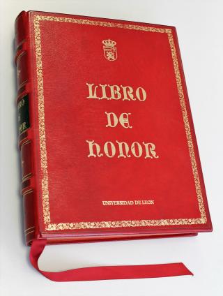Fotografía de un libro de tapa dura color rojo, con detalles en relieve dorado. En la portada se lee en letras grandes y doradas: 'LIBRO DE HONOR'. Sobre el título hay un escudo pequeño también en dorado. El libro tiene un marcador de cinta rojo sobresaliendo de la parte inferior y un lomo con decoraciones doradas y una etiqueta negra.