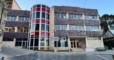“Edificio del Ayuntamiento de Alcañiz visto desde la calle, con tres plantas, fachada de piedra oscura y grandes ventanas. Destaca una torre acristalada cilíndrica con franjas rojas y blancas. En la parte inferior se observa la entrada principal y una calle adoquinada frente al edificio.”