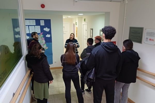 Grupo de personas reunidas en un pasillo interior, escuchando a una mujer que parece estar explicando algo durante una visita guiada o actividad educativa. En las paredes hay carteles y decoración escolar.