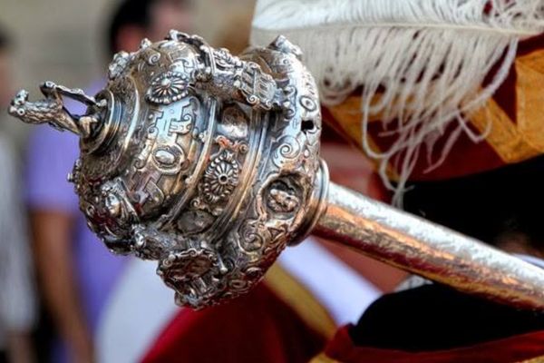 Maza ceremonial de metal plateado con relieves ornamentales detallados, sostenida por una persona vestida con indumentaria tradicional roja y dorada, con una pluma blanca visible en el atuendo.