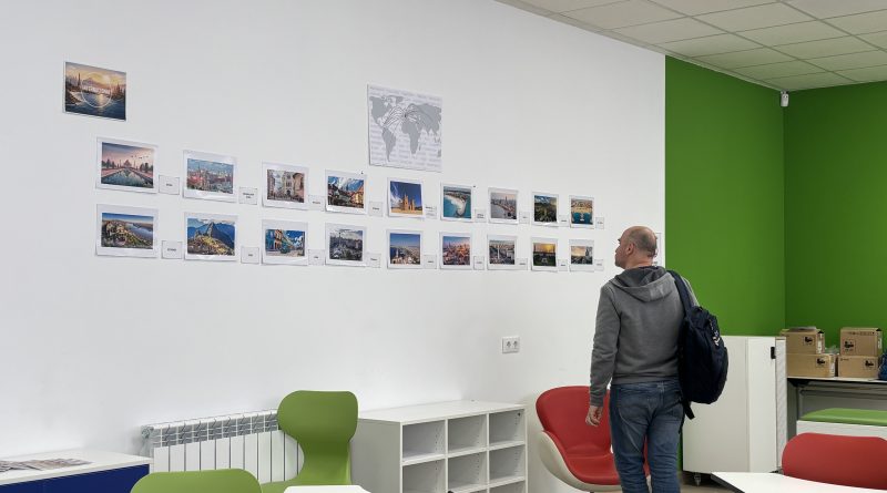 Persona observando una exposición de fotografías de diferentes ciudades y países colocadas en una pared blanca en un espacio del centro educativo, durante una muestra sobre los orígenes culturales del alumnado y profesorado.
